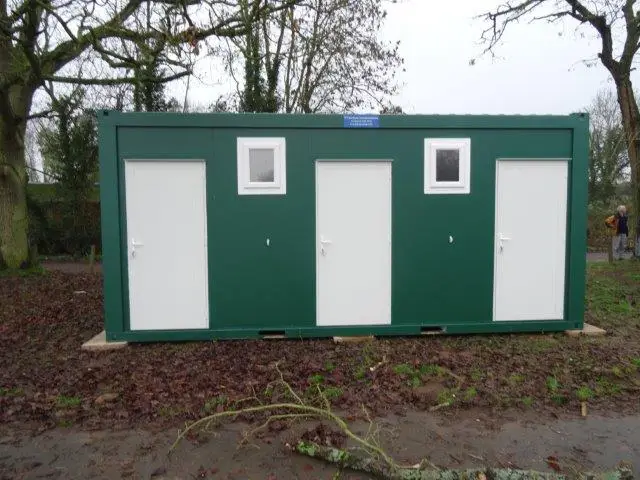 Green modular building with three doors and windows in a leafy setting.