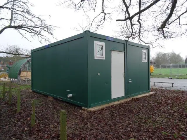Rear view of a green modular building set against a backdrop of trees in a park.