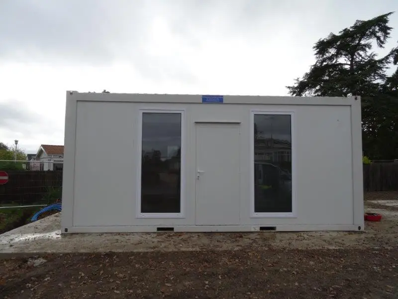 Exterior of a white modular building with glass doors and a flat roof, set on a muddy construction site
