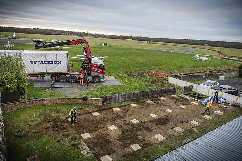 A crane lifting a portable building onto a prepared base on a construction site, with a worker guiding the placement.