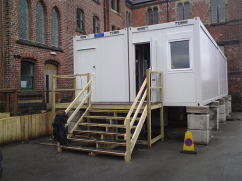 Portable modular building with stairs and railing installed beside an old brick building, possibly for temporary office or catering use