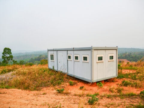 Modular portable building positioned in a remote, grassy area with a hazy forested landscape in the background