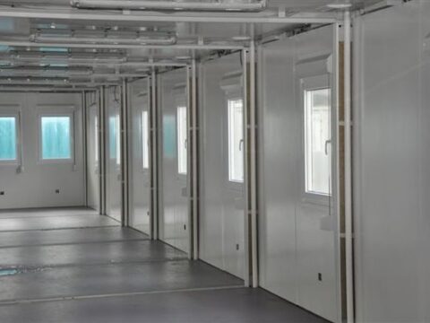 Open-plan modular building interior with exposed ceiling infrastructure, multiple doors, and windows allowing for natural light